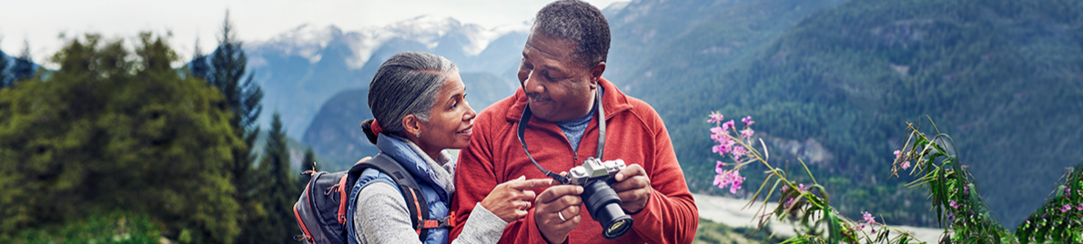 With wooded mountains in the distance, a couple smiles at each
                      other while looking at photos on a camera.